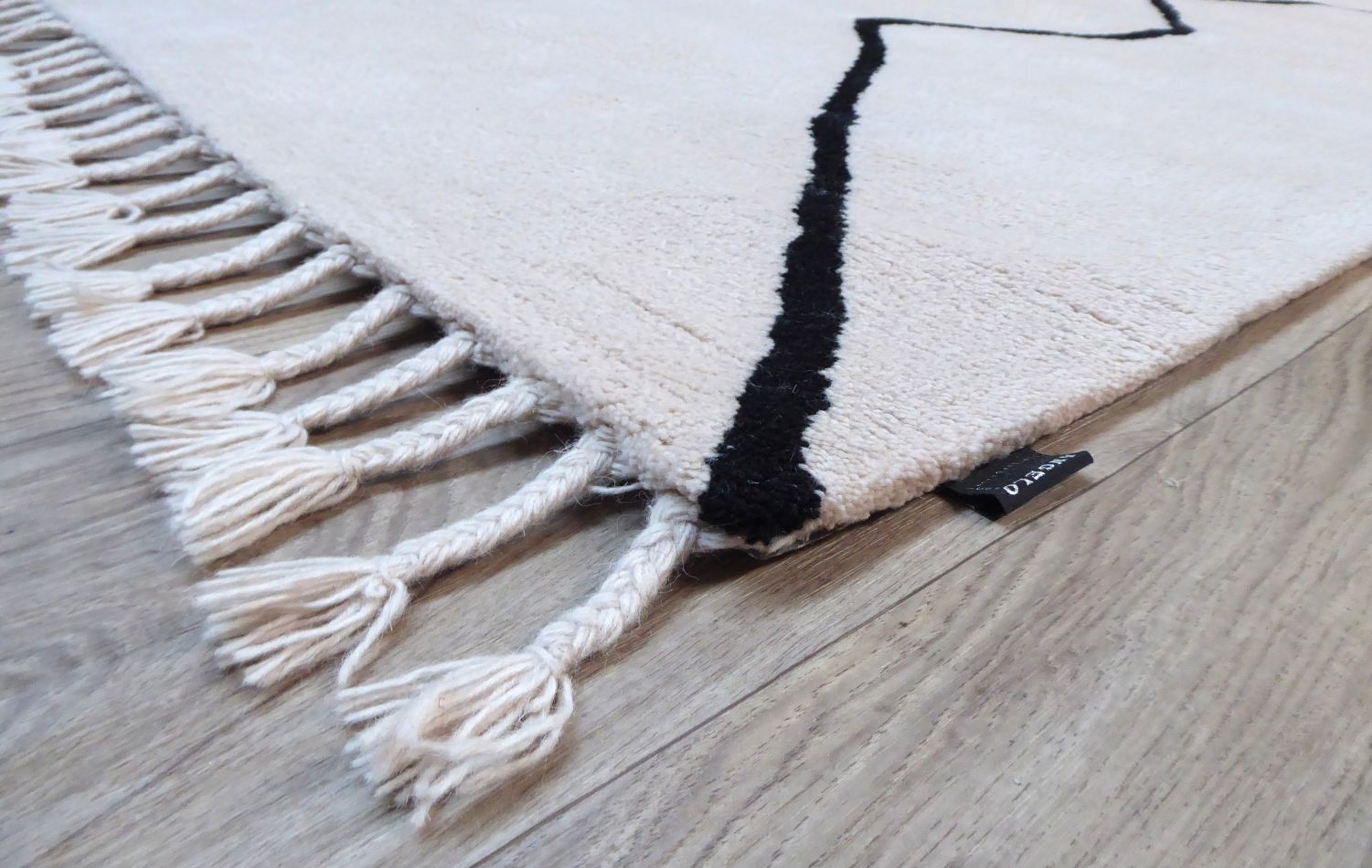 Détail d’un coin du tapis ivoire en laine avec franges et motif noir discret